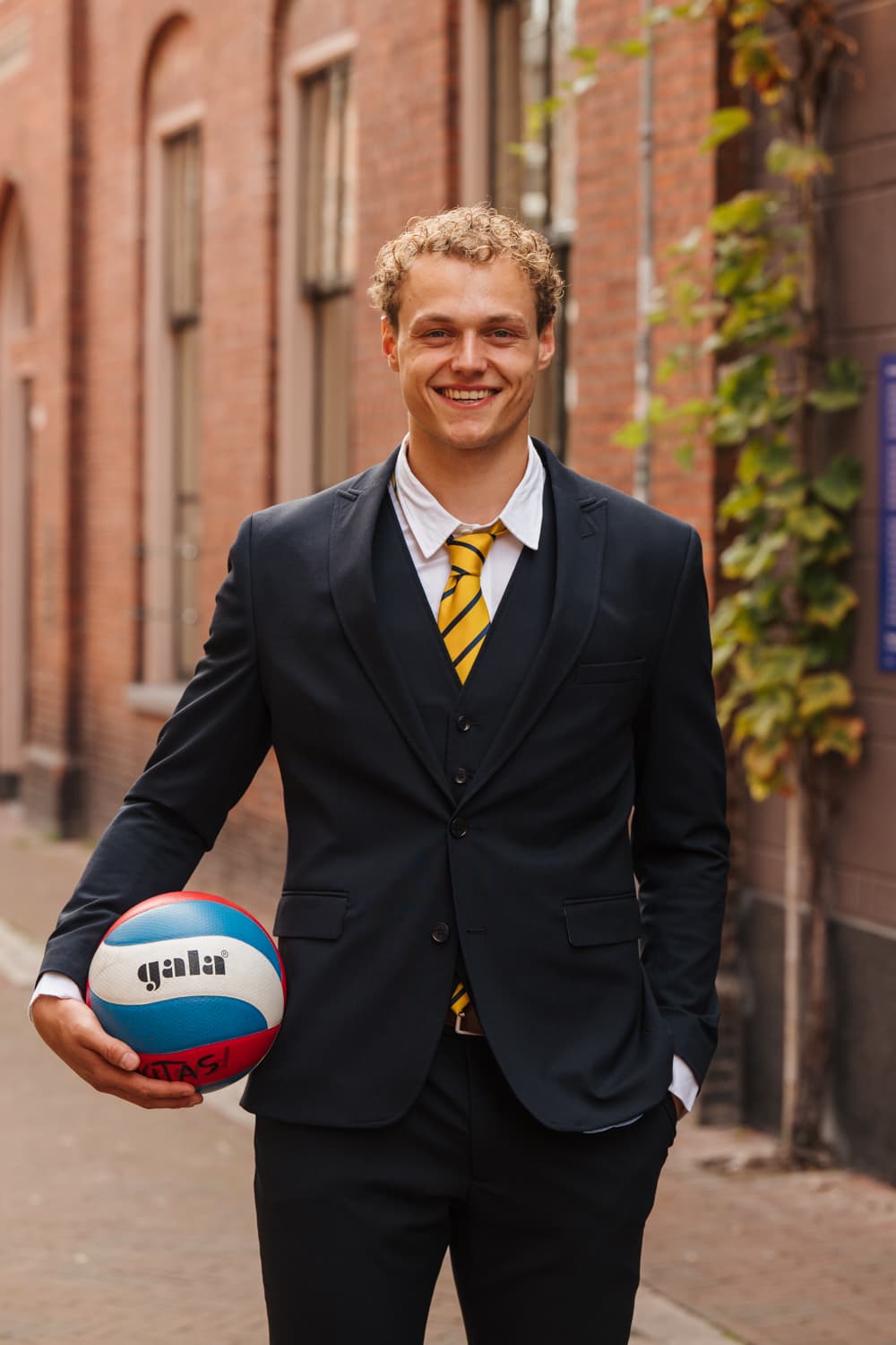 Tjalle-Durk van der Eems in pak met een volleybal onder de arm, voor een bakstenen gevel